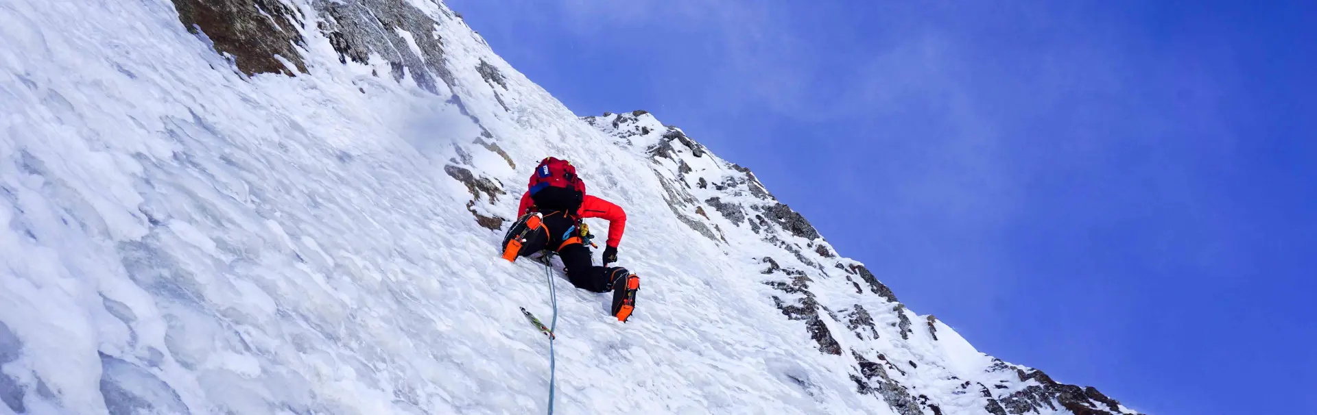 Een bergbeklimmer in rode winterkleding zit op een besneeuwde bergkam, met uitgestrekte armen en stokken in de hand, tegen een achtergrond van een indrukwekkende overhangende sneeuwformatie en een bew